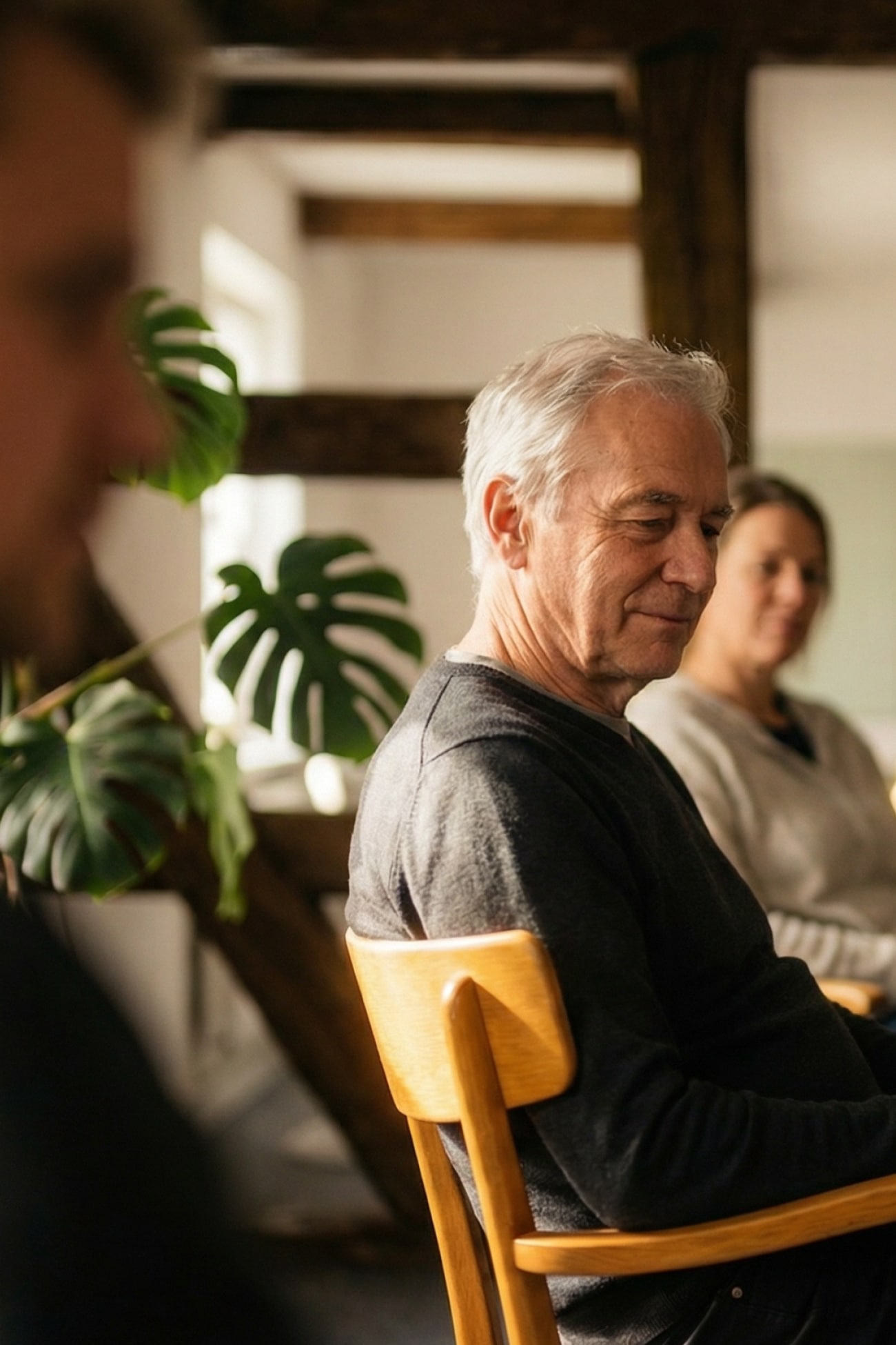 Eine Gruppe von Menschen sitzt in einem hellen Raum, mit Pflanzen im Hintergrund. Fokus auf einen älteren Mann.