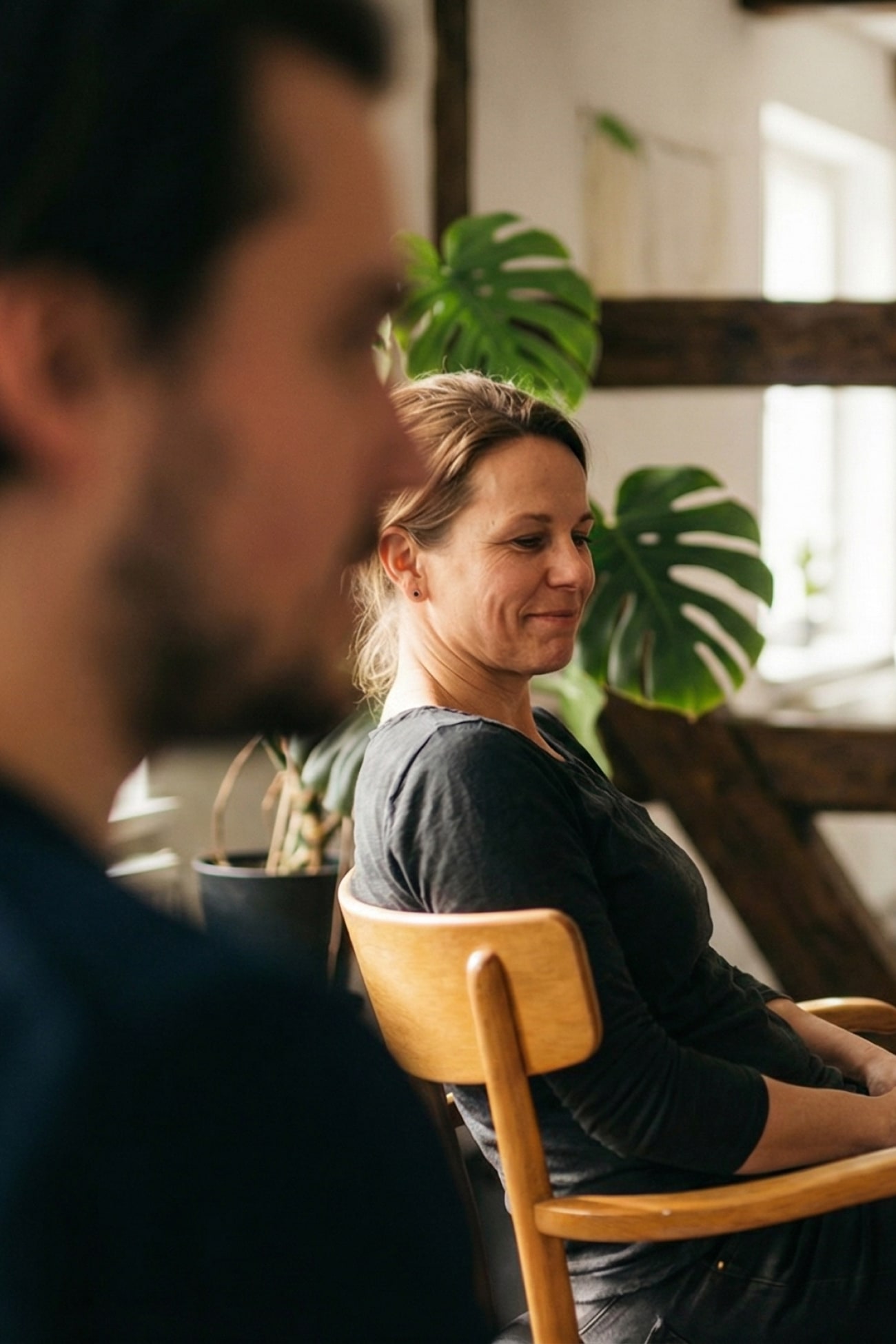 Zwei Personen in einem Raum, eine Frau sitzt lächelnd auf einem Stuhl, im Hintergrund eine Pflanze.