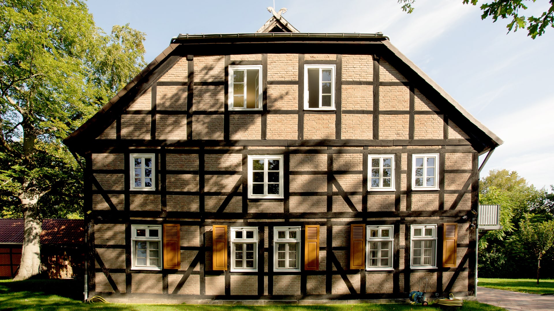 Fachwerkhaus mit weißen Fenstern und Holzläden, umgeben von Bäumen und Rasen.