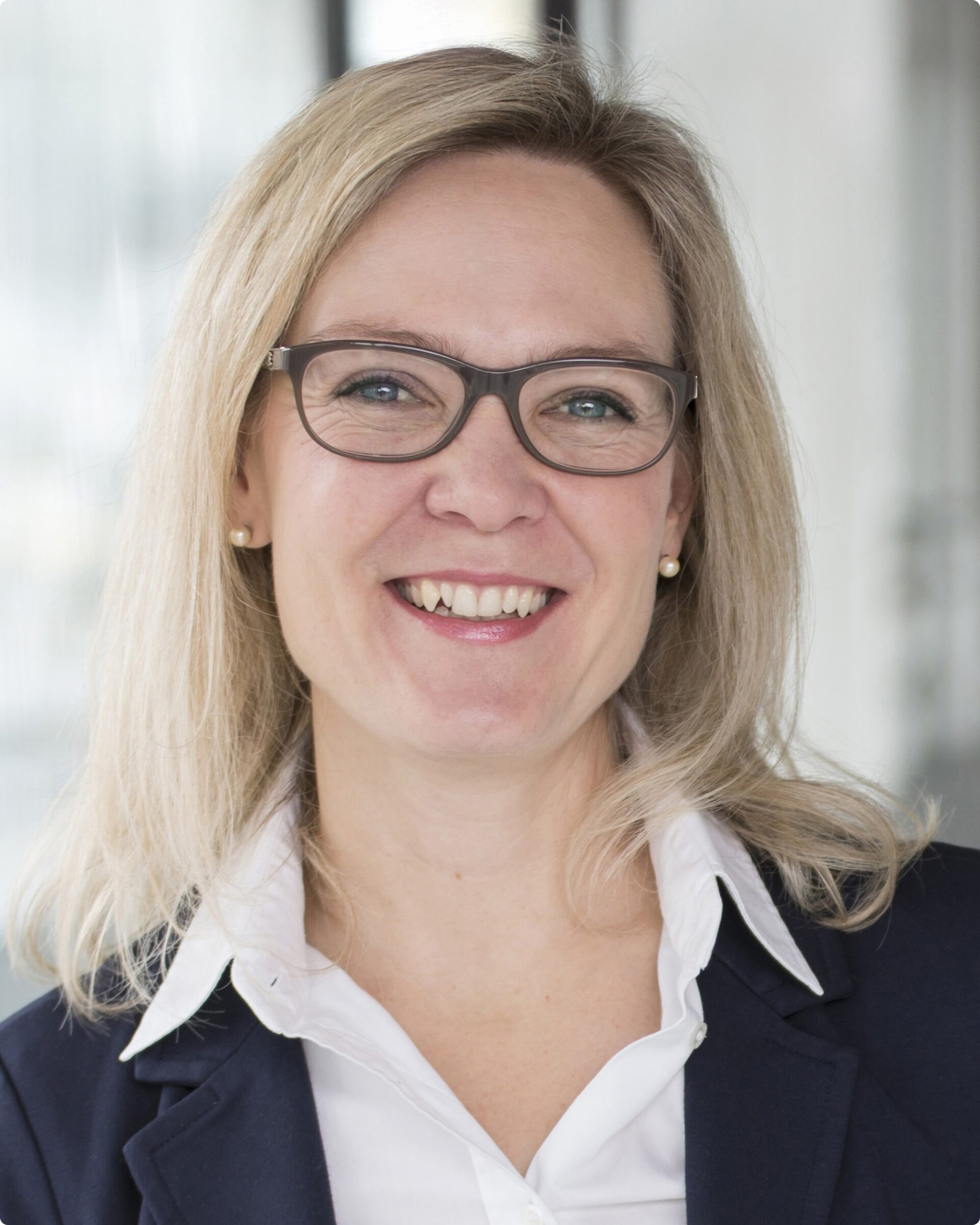 Portrait einer lächelnden Frau mit blonden Haaren, Brille und einem blauen Blazer, vor einem modernen Hintergrund.