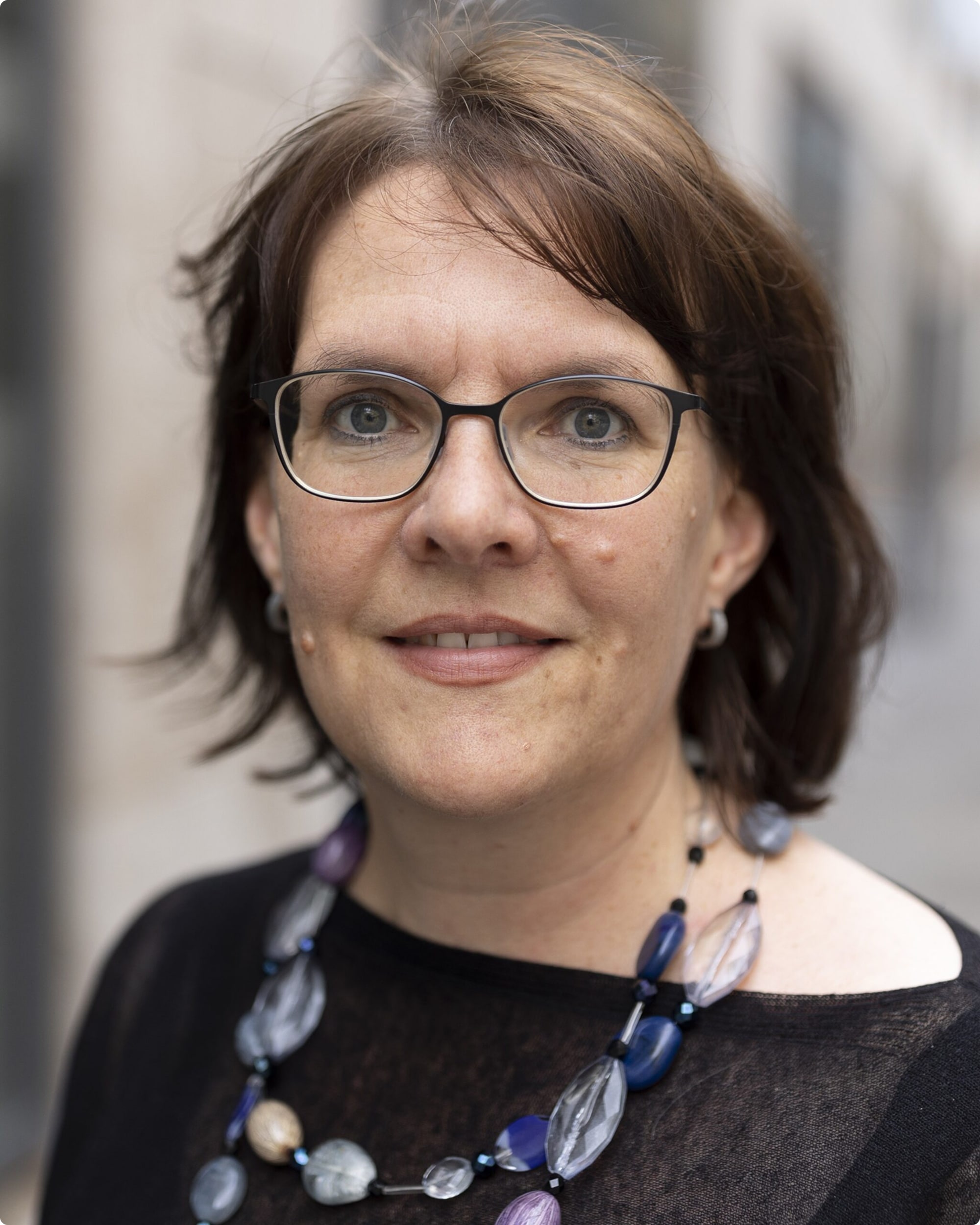 Porträt einer Frau mit Brille und Halskette, lächelnd vor unscharfem Hintergrund.