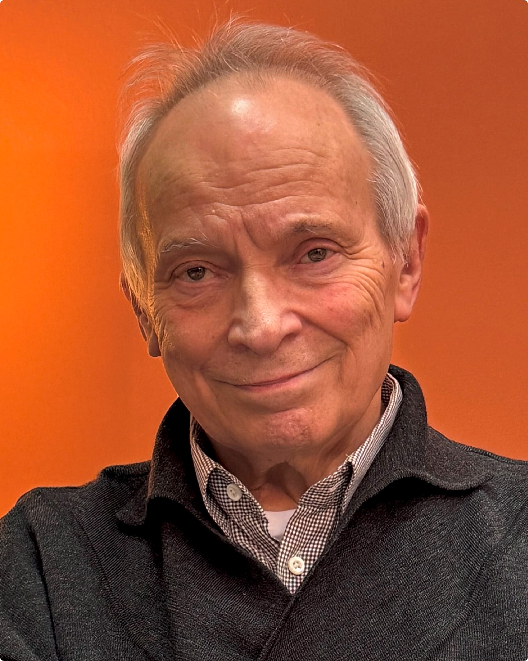 Porträt eines älteren Mannes mit grauen Haaren, lächelnd vor orangefarbenem Hintergrund.