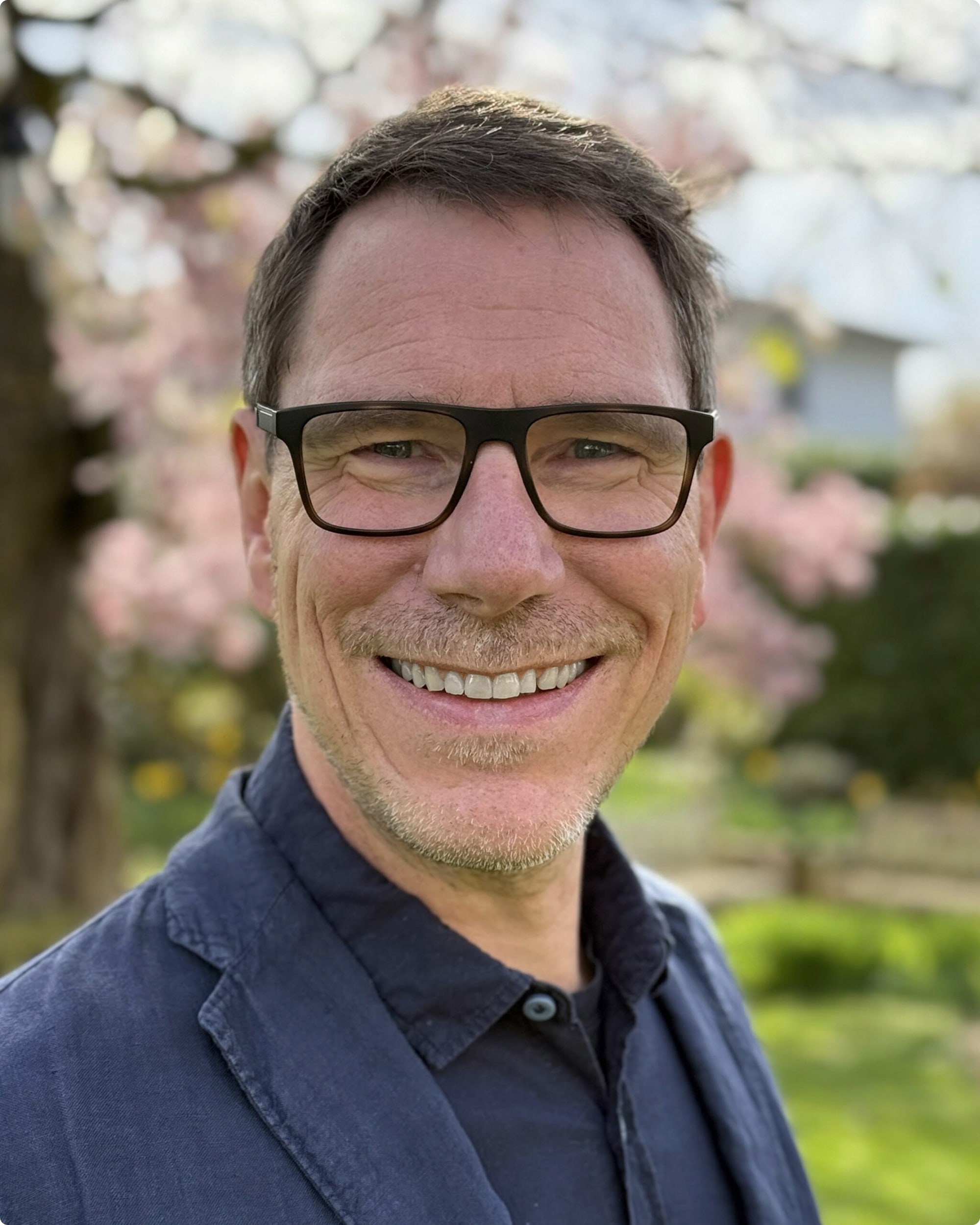 Porträt eines lächelnden Mannes mit Brille, im Freien vor blühenden Bäumen.