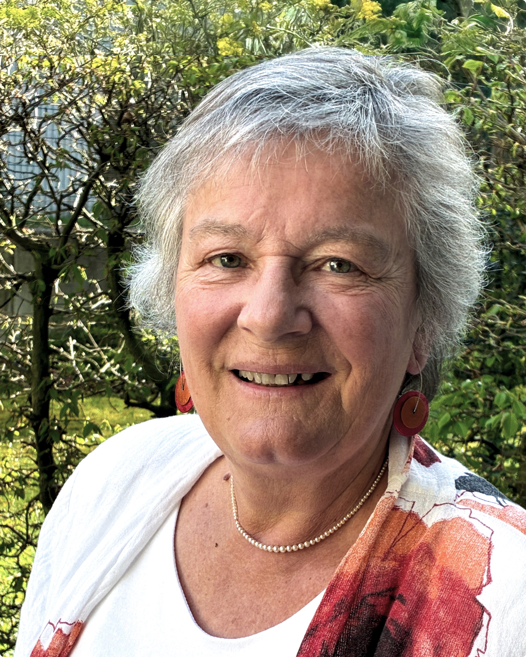 Porträt einer lächelnden älteren Frau mit grauen Haaren, die eine bunte Bluse und Schmuck trägt, vor einem grünen Hintergrund