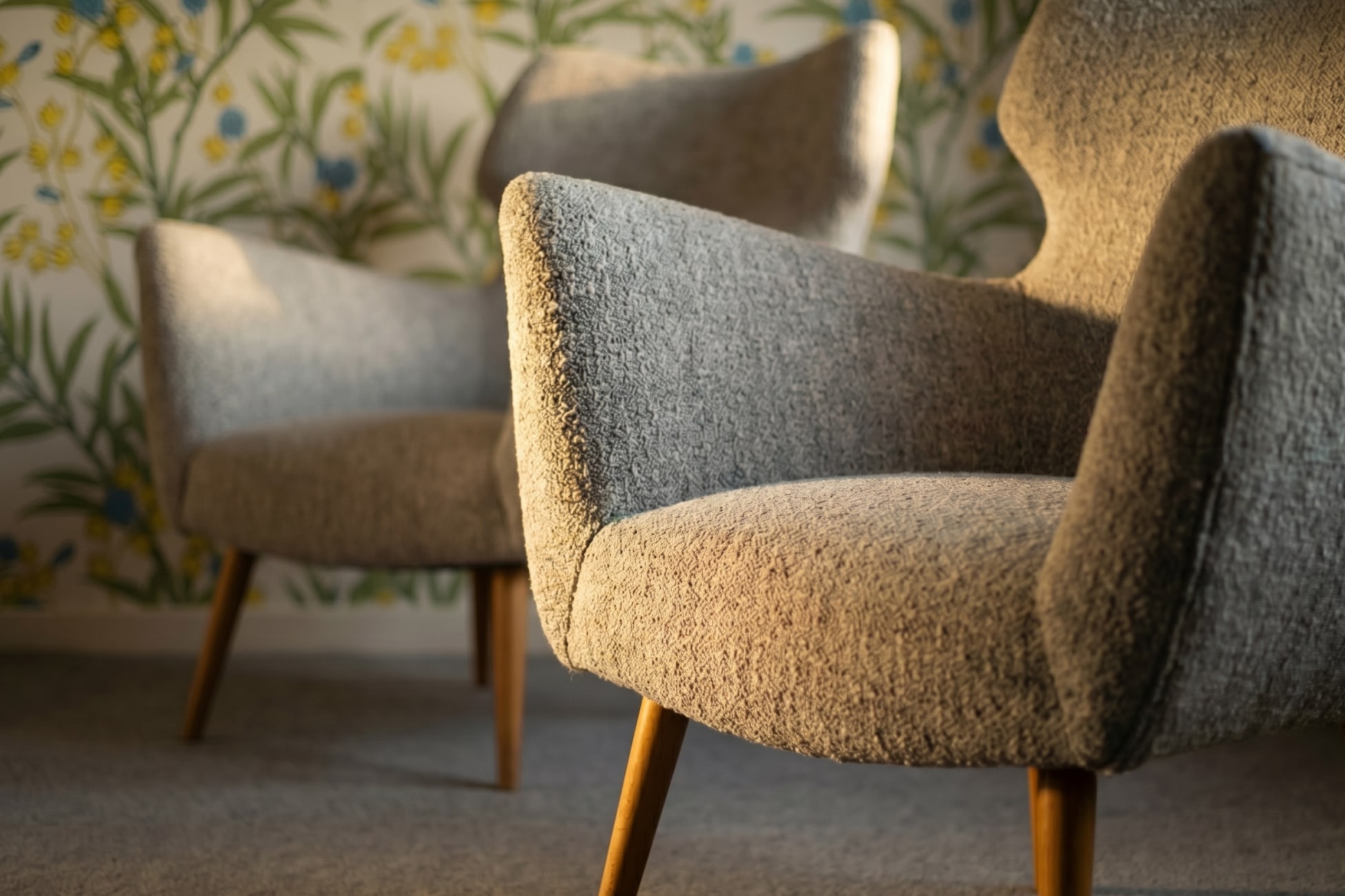 Zwei moderne Sessel mit Holzbeinen vor einer blumigen Tapete in sanften Farben.